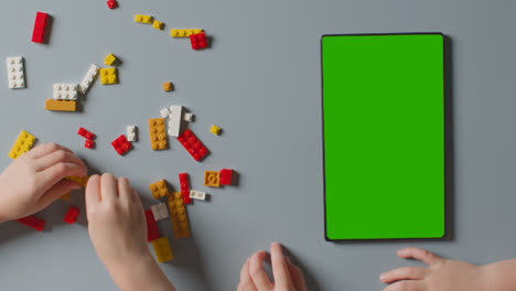 Draufsicht-Auf-Zwei-Kinder,-Die-Mit-Plastikbausteinen-Neben-Einem-Digitalen-Tablet-Mit-Grünem-Bildschirm-Spielen